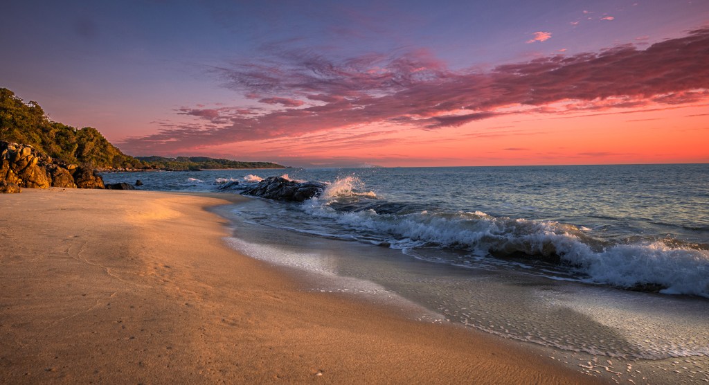 A serene beach scene at sunset, featuring soft sands, gentle waves, and a colorful sky filled with pink and purple hues.