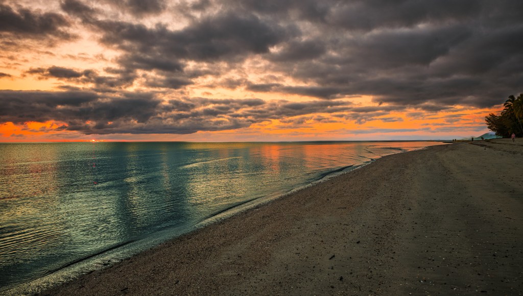Scenic beach view at sunset with a calm sea and dramatic clouds reflecting colors in the water.
