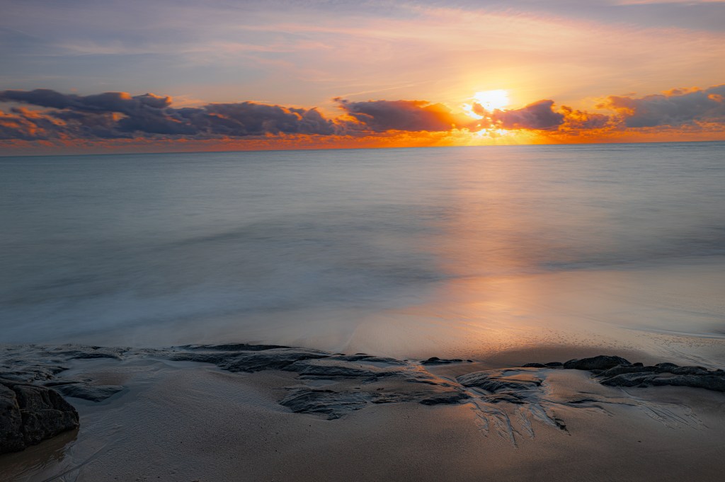 A serene beach scene at sunset, showcasing a vibrant sky with orange and yellow hues, gentle waves reflecting the sunset, and rocky shoreline in the foreground.