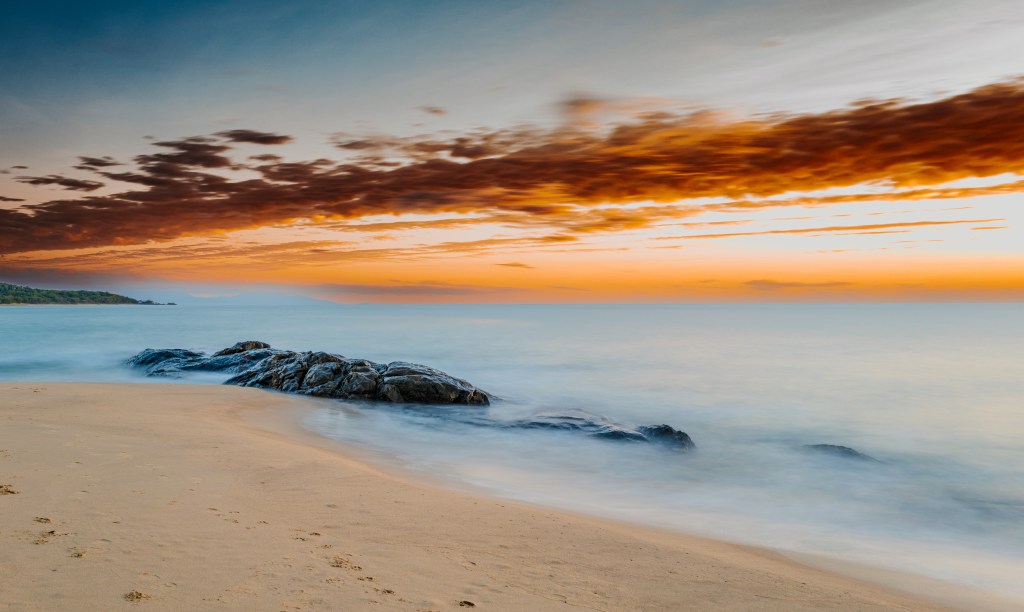 A serene beach scene at sunset, featuring smooth sand, a rocky formation near the water's edge, and a colorful sky with orange and blue hues.